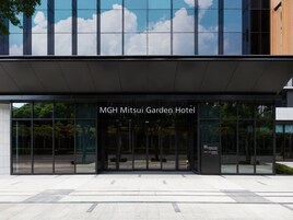 MGH Mitsui Garden -hotellin sisäänkäynti, jossa on moderni lasijulkisivu ja josta avautuu esteetön näkymä ympäröivään vihreään maisemaan.