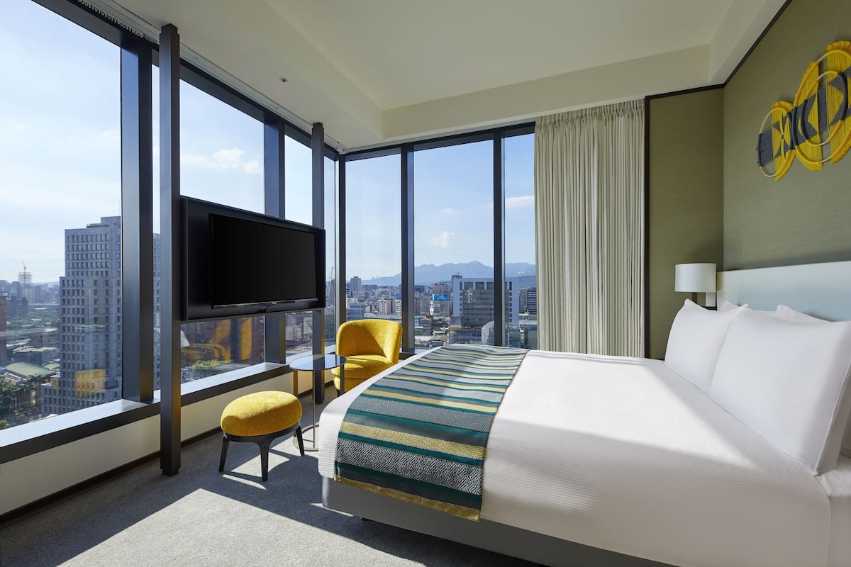 A hotel room with a large bed, a TV mounted on the wall, a yellow chair, and a view of the city through large windows.