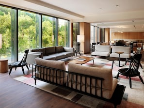 A modern living room with a sofa, chairs, and a coffee table. Large windows offer a view of greenery outside.