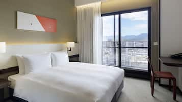 A hotel room with a large bed, a desk, a chair, and a view of the city through a large window.