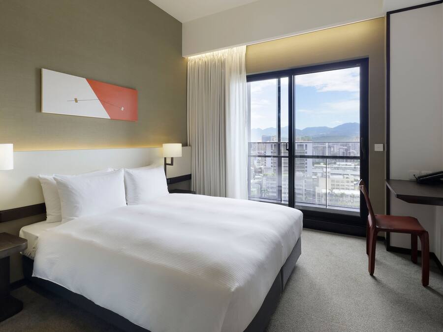 A hotel room with a large bed, a desk, a chair, and a view of the city through a large window.