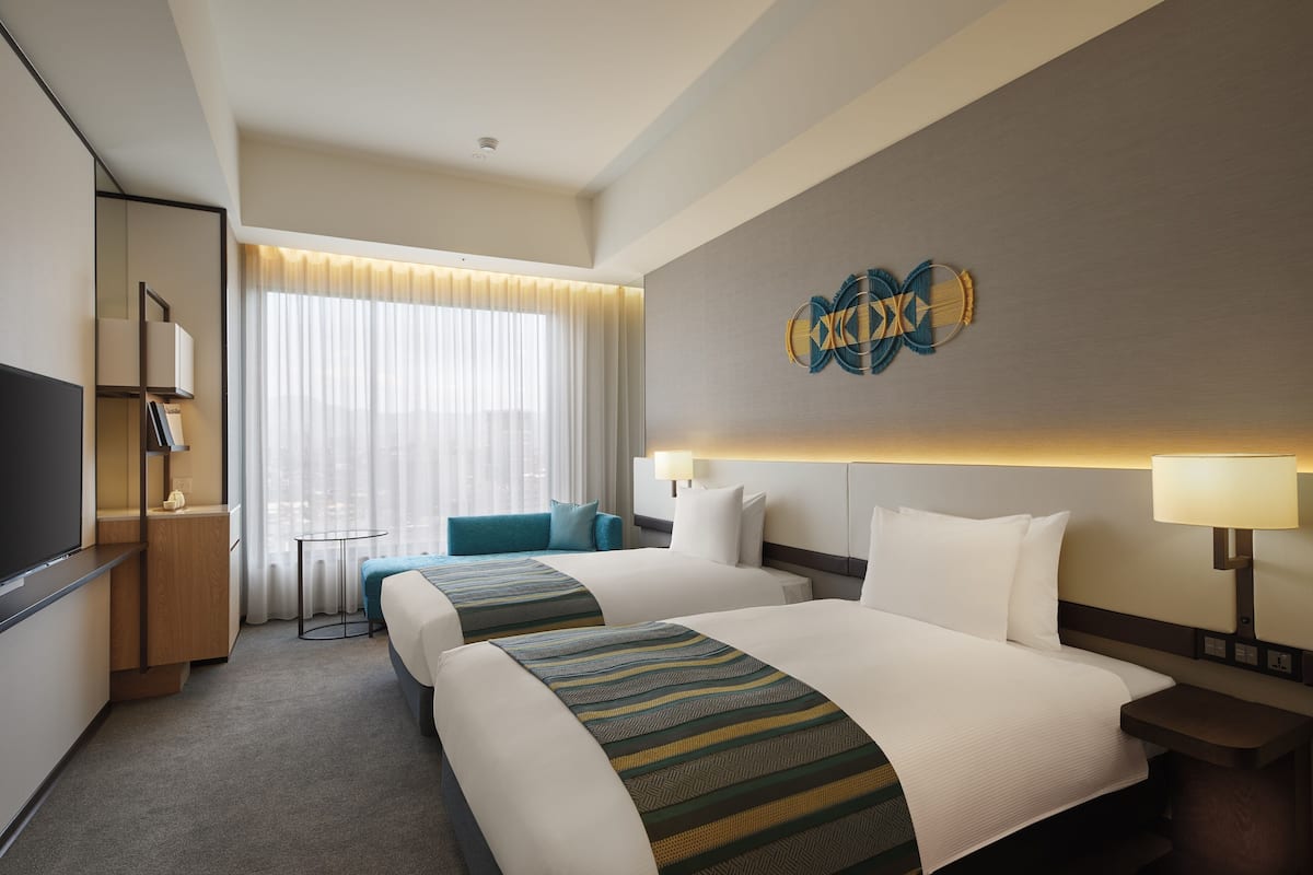 A hotel room with two beds, a TV, a desk, and a window with curtains.
