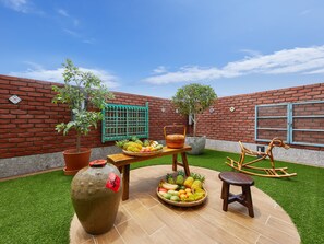 Un patio con pared de ladrillo, césped artificial verde, una mesa de madera con cestas de fruta, una mecedora y plantas en macetas.