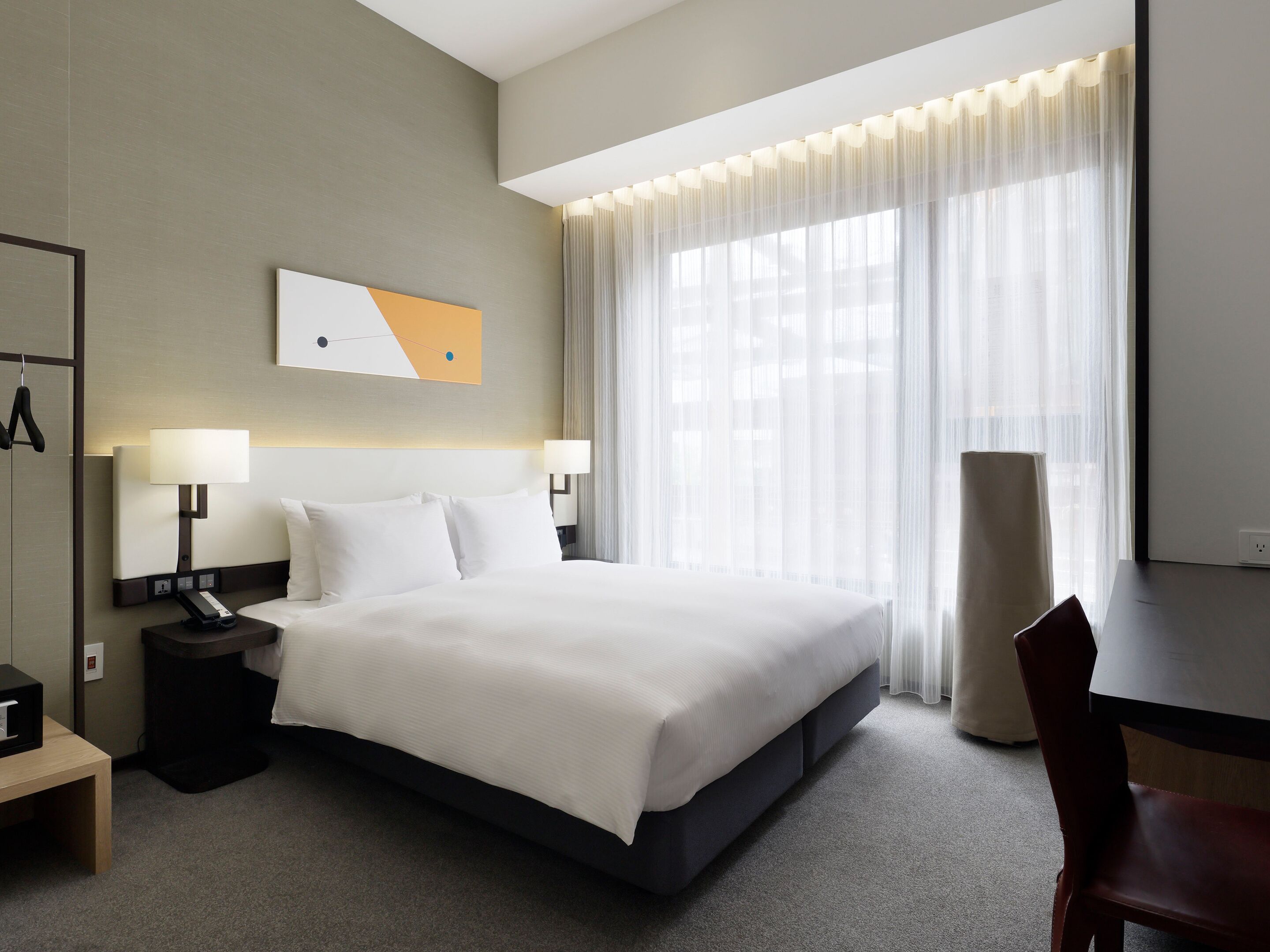 A hotel room with a large bed, bedside lamps, a desk, a chair, and a window with sheer curtains.