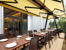 An outdoor dining area with multiple tables and chairs, a yellow awning, and a view of the interior through glass doors.