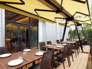 An outdoor dining area with multiple tables and chairs, a yellow awning, and a view of the interior through glass doors.