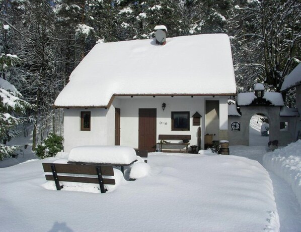Un pequeño edificio cubierto de nieve con un banco de madera al frente, rodeado de árboles.