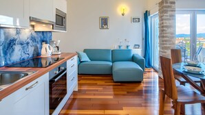 A modern kitchen with a marble backsplash, a built-in oven, and a coffee maker. There is a blue sofa and a dining table with chairs. The room has a large window with a view of the city. - Modish Apartment in Murter with Hot Tub (Murter-Kornati)