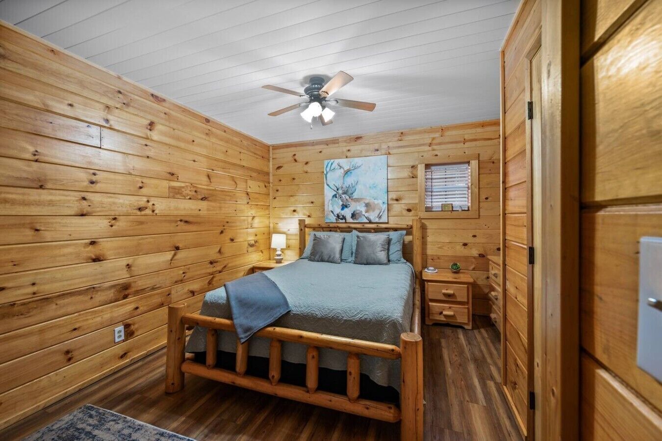 A wooden cabin bedroom with a bed, nightstands, a ceiling fan, and a painting of deer.