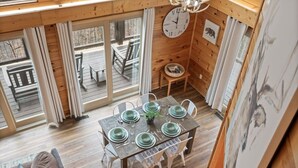 Un intérieur en bois avec une table à manger dressée pour quatre personnes, une horloge au mur et une vue dégagée sur l’extérieur grâce à de grandes fenêtres.