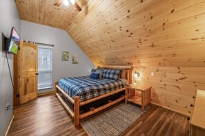 Un dormitorio acogedor con techo de madera, una cama con ropa de cama a cuadros, una mesita de noche con una lámpara y un televisor montado en la pared.