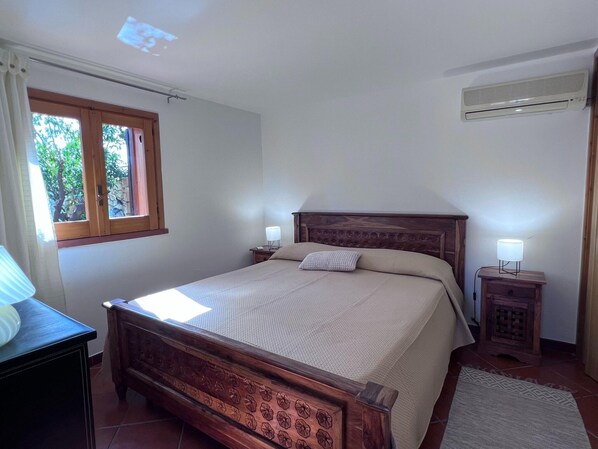 A bedroom with a wooden bed, two bedside lamps, a window with curtains, an air conditioner, and a tiled floor. - Charming Holiday Home Near Sea (Castellammare del Golfo)