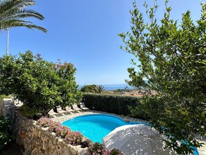 Ein Pool mit Steinmauer, umgeben von viel Grün und Blick auf das Meer.