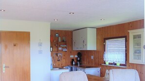 Une cuisine compacte avec des meubles en bois, un évier et une fenêtre avec des stores. On y trouve deux chaises avec des coussins et une petite table.