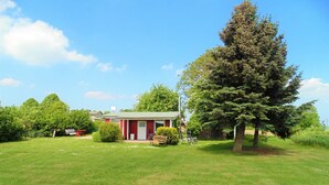 Une maison rouge avec une porte blanche et un petit porche, entourée d’une pelouse bien entretenue et d’un grand arbre à feuilles persistantes.