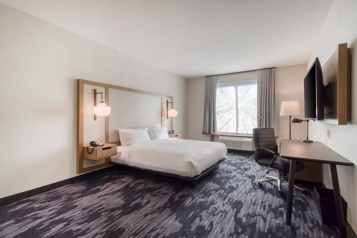 A hotel room with a bed, bedside table, lamp, desk, chair, and a TV mounted on the wall.