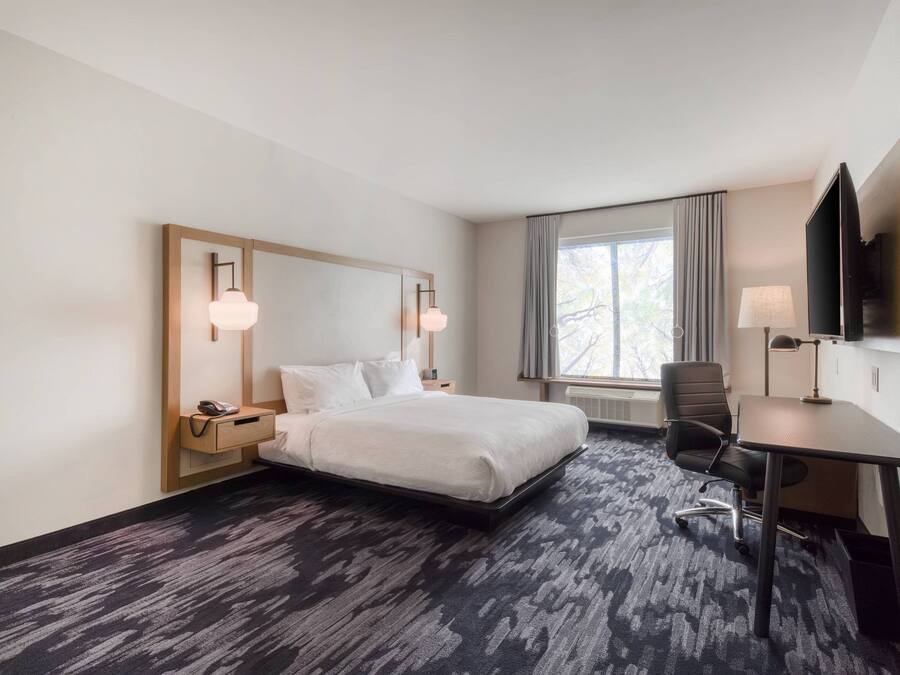 A hotel room with a bed, bedside table, lamp, desk, chair, and a TV mounted on the wall.