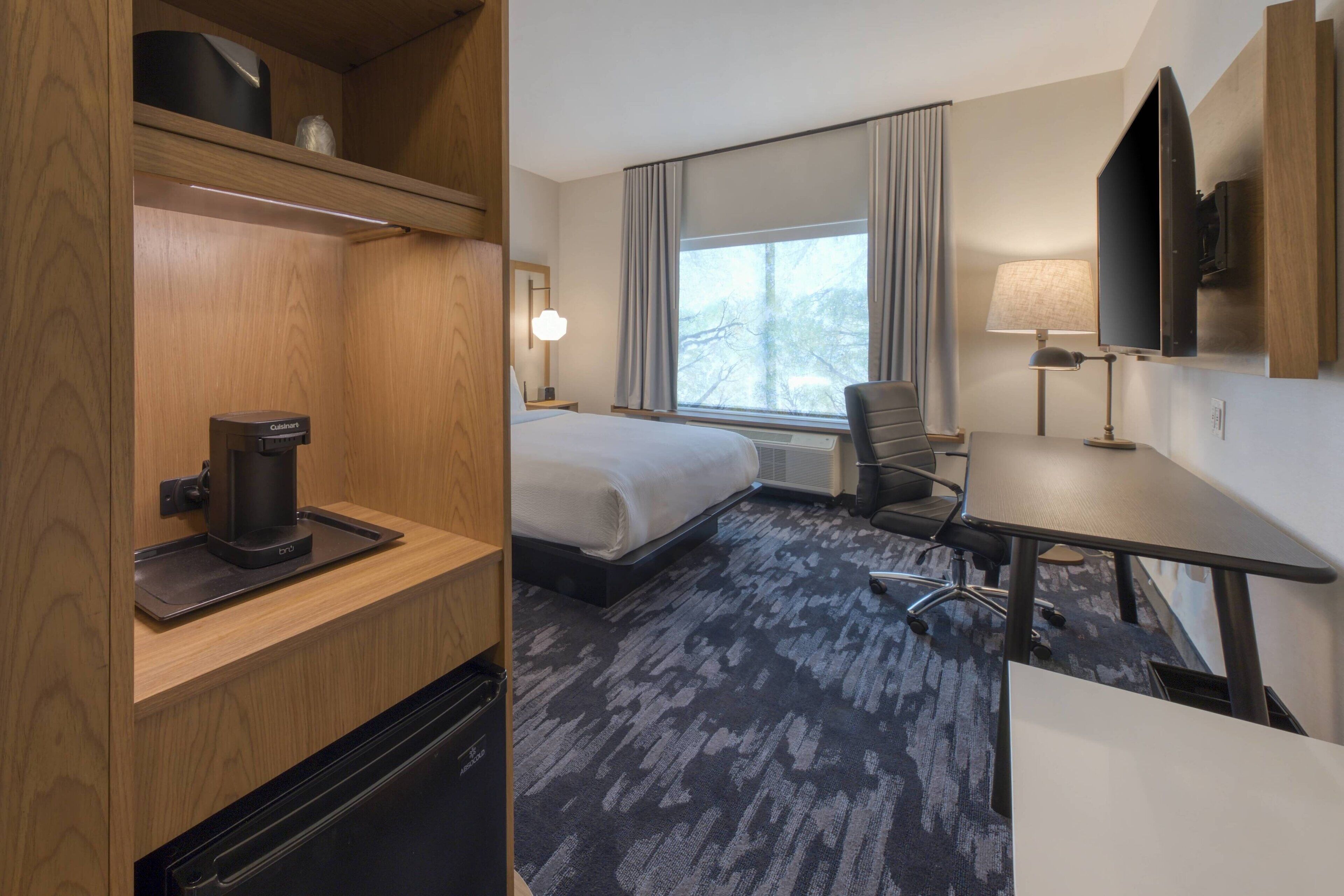 Habitación de hotel con cama, escritorio, silla, cafetera, televisión y ventana con cortinas.