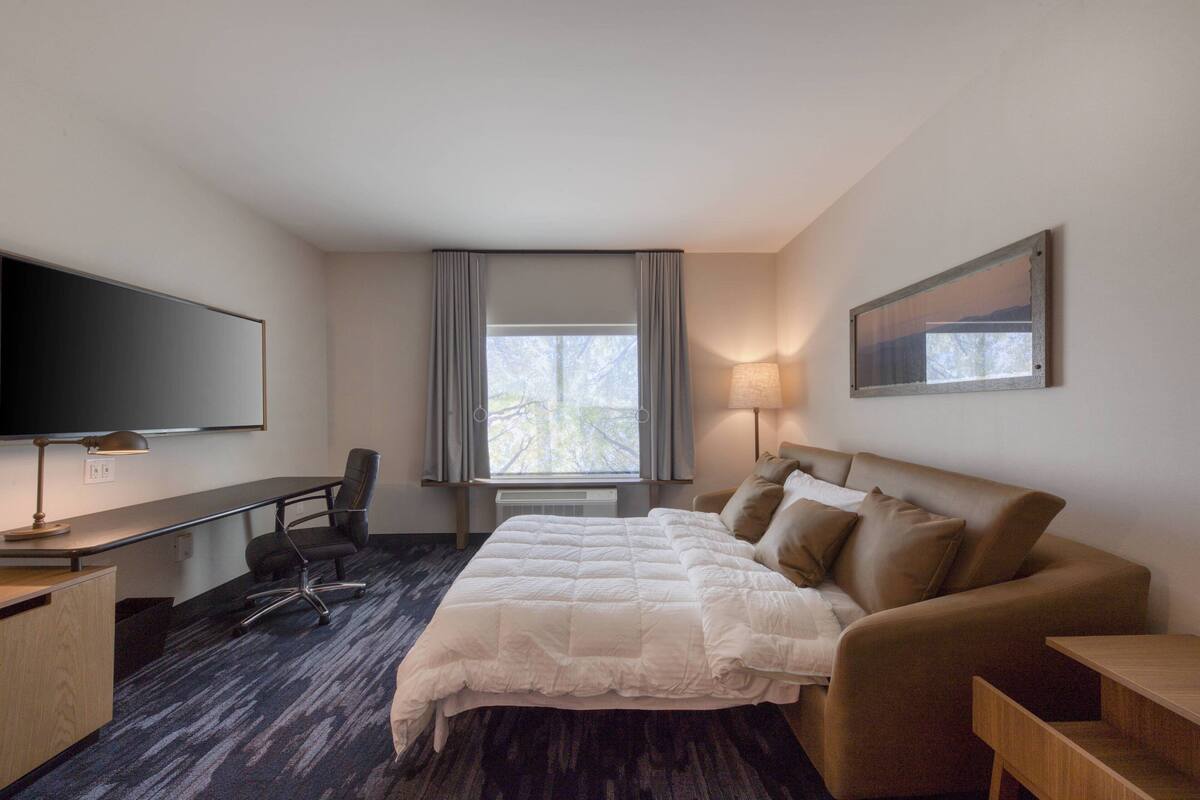 A hotel room with a bed, a desk with a chair, a TV, a window with curtains, and a lamp.