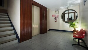 Un couloir avec un escalier, une porte en bois, une chaise rouge, une plante en pot et un extincteur.