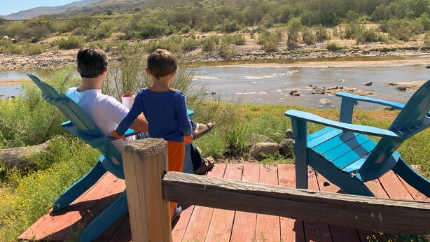 Une terrasse en bois avec deux chaises bleues donnant sur une rivière et des montagnes.
