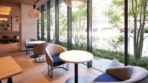 Decoração moderna com mesas redondas e cadeiras estofadas, amplas janelas com vista para áreas verdes e edifícios, e iluminação pendente.