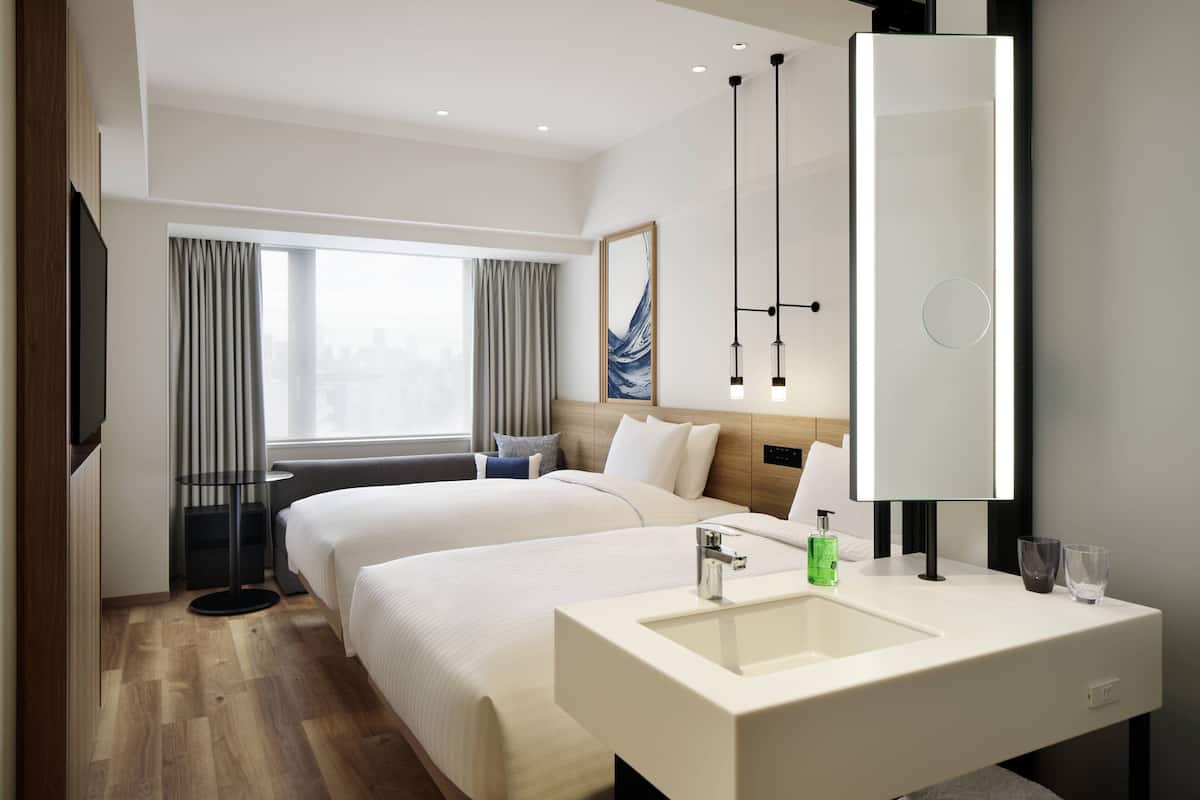 A modern hotel room with two beds, a large mirror, a white sink, and a city view through the window.