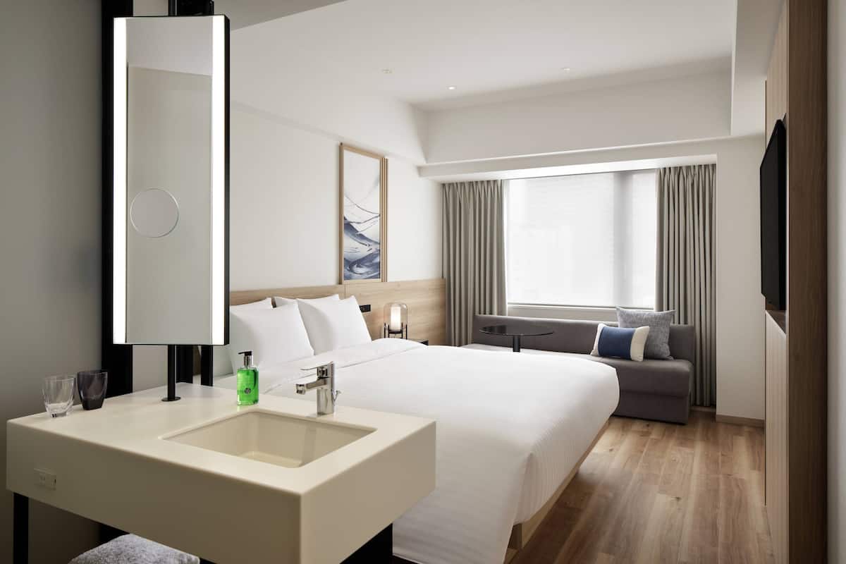 A hotel room with a large bed, a sofa, a TV, a mirror, and a bathroom with a sink and two glasses.