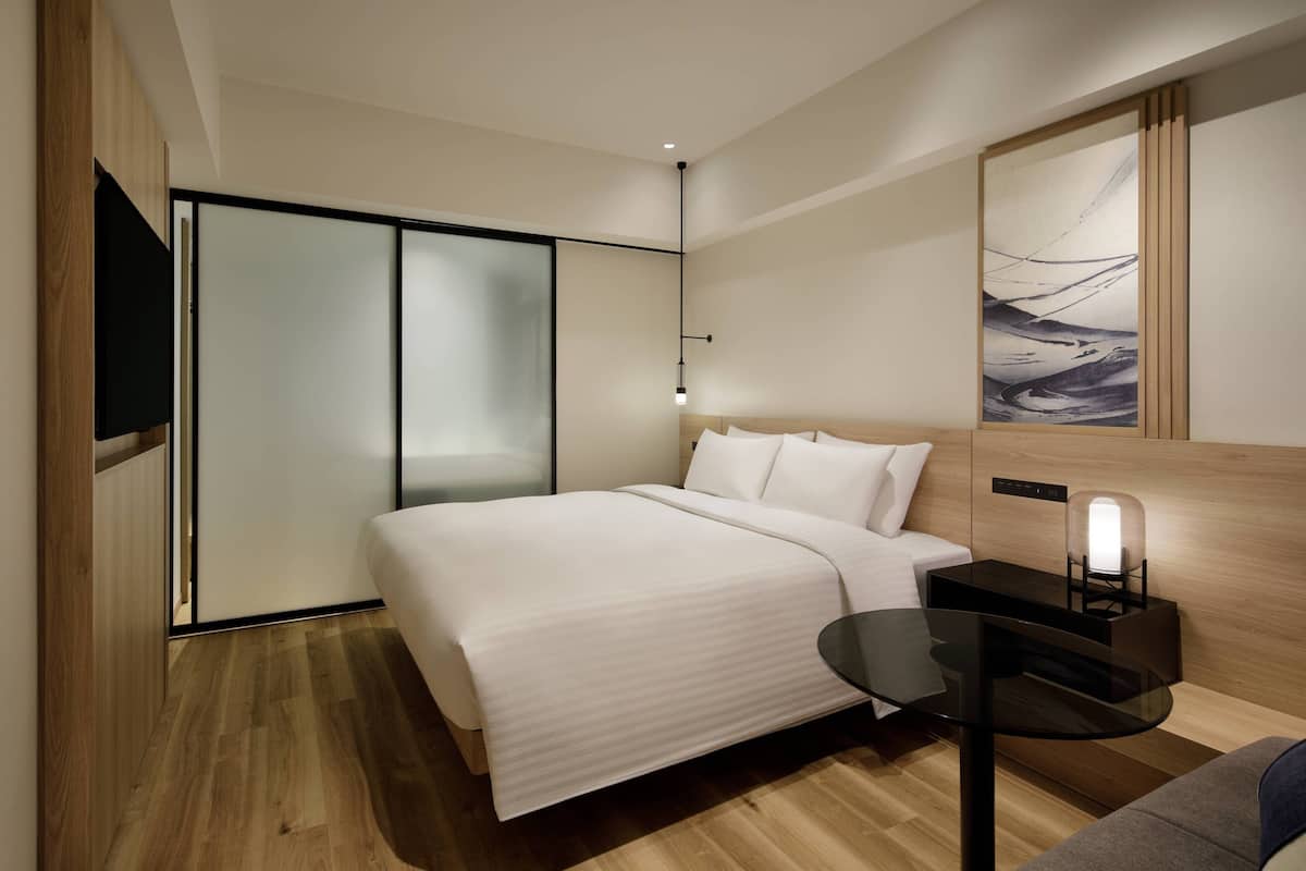 A modern hotel room with a large bed, a bedside table with a lamp, a wall-mounted TV, and a sliding door closet.