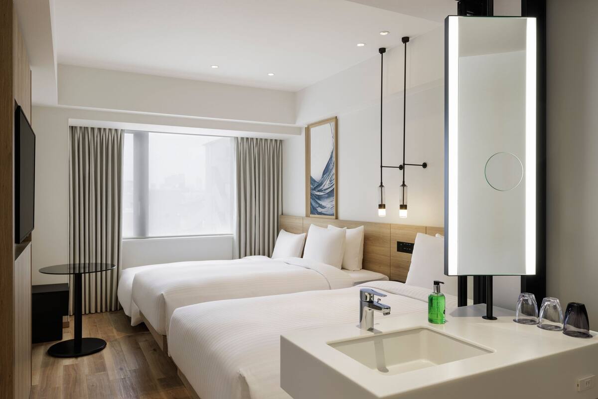 A hotel room with two beds, a large mirror, a round table, and a modern bathroom with a sink and glassware.