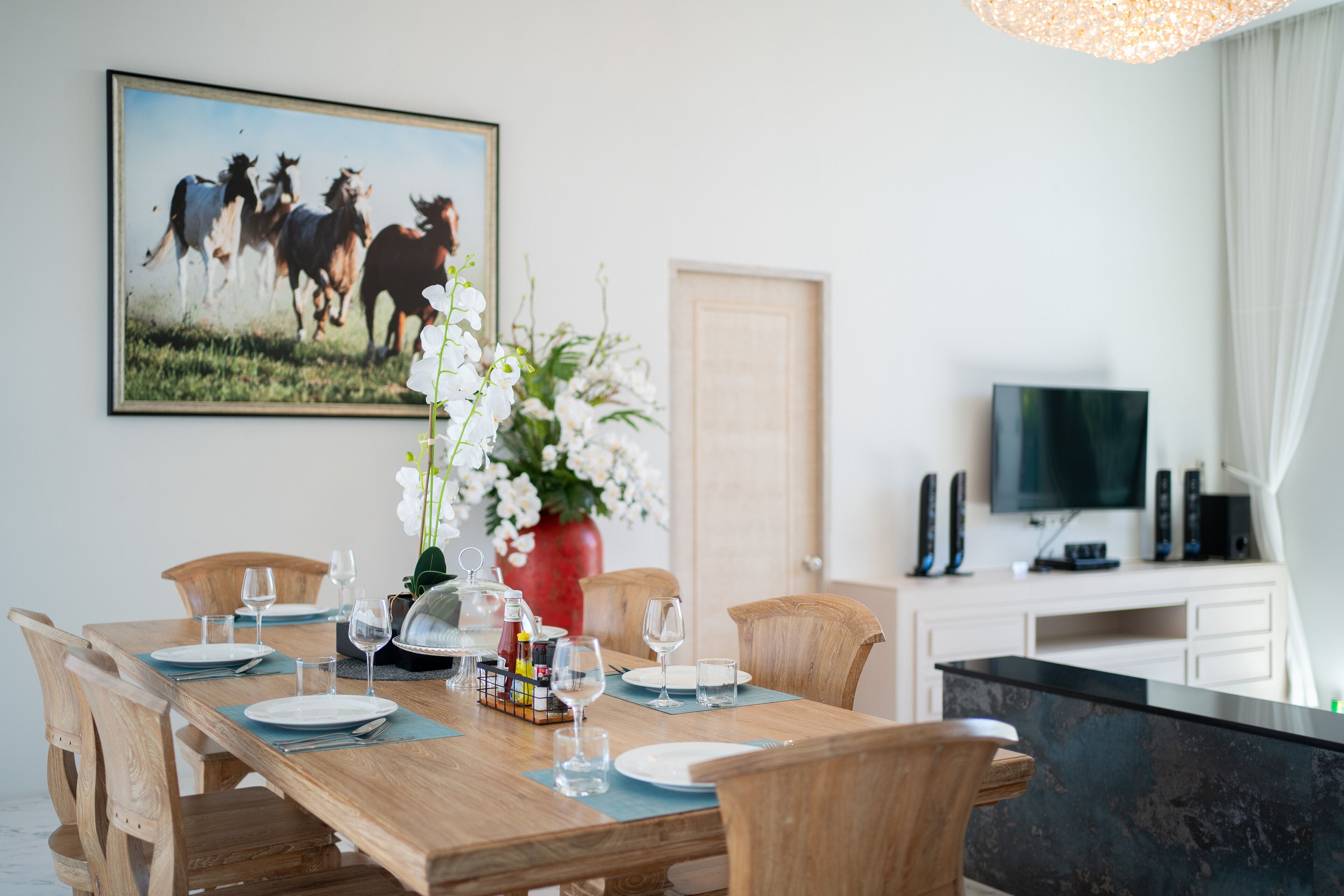 A dining area with a wooden table set for six, surrounded by wooden chairs. A painting of horses is on the wall, and a television is mounted on the wall to the right.