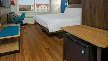 Habitación de hotel con cama, escritorio, silla, refrigerador pequeño y ventana con vista a edificios.
