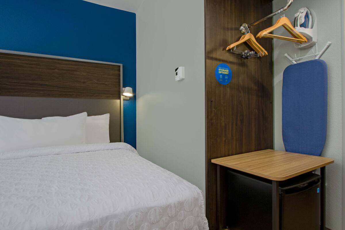 Hotellihuone, jossa on sänky, pieni pöytä, tuoli, naulakko ja kyltti, jossa kerrotaan säilytysmahdollisuuksista.