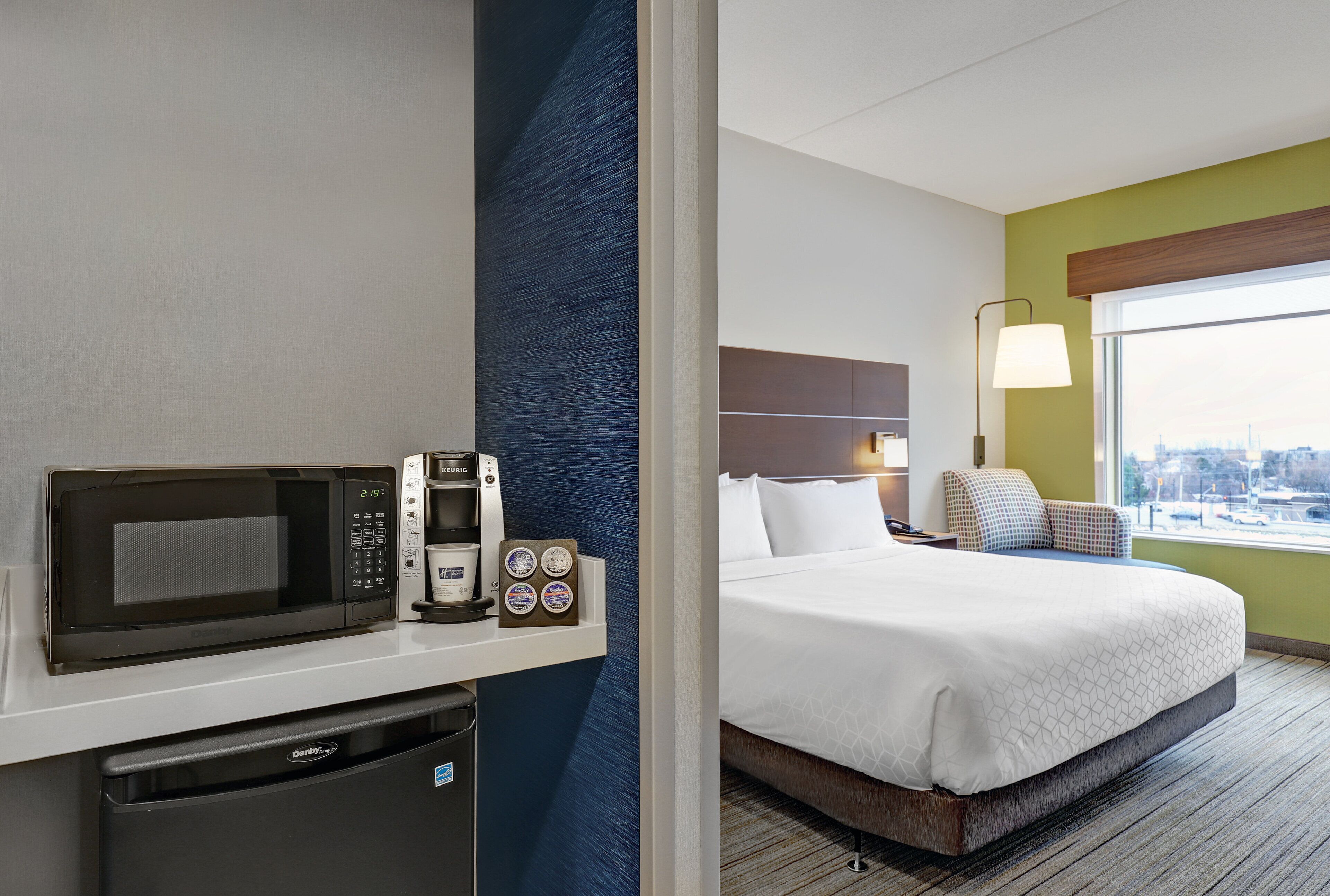 Een hotelkamer met een bed, een magnetron, een koffiezetapparaat en uitzicht op de stad door het raam.