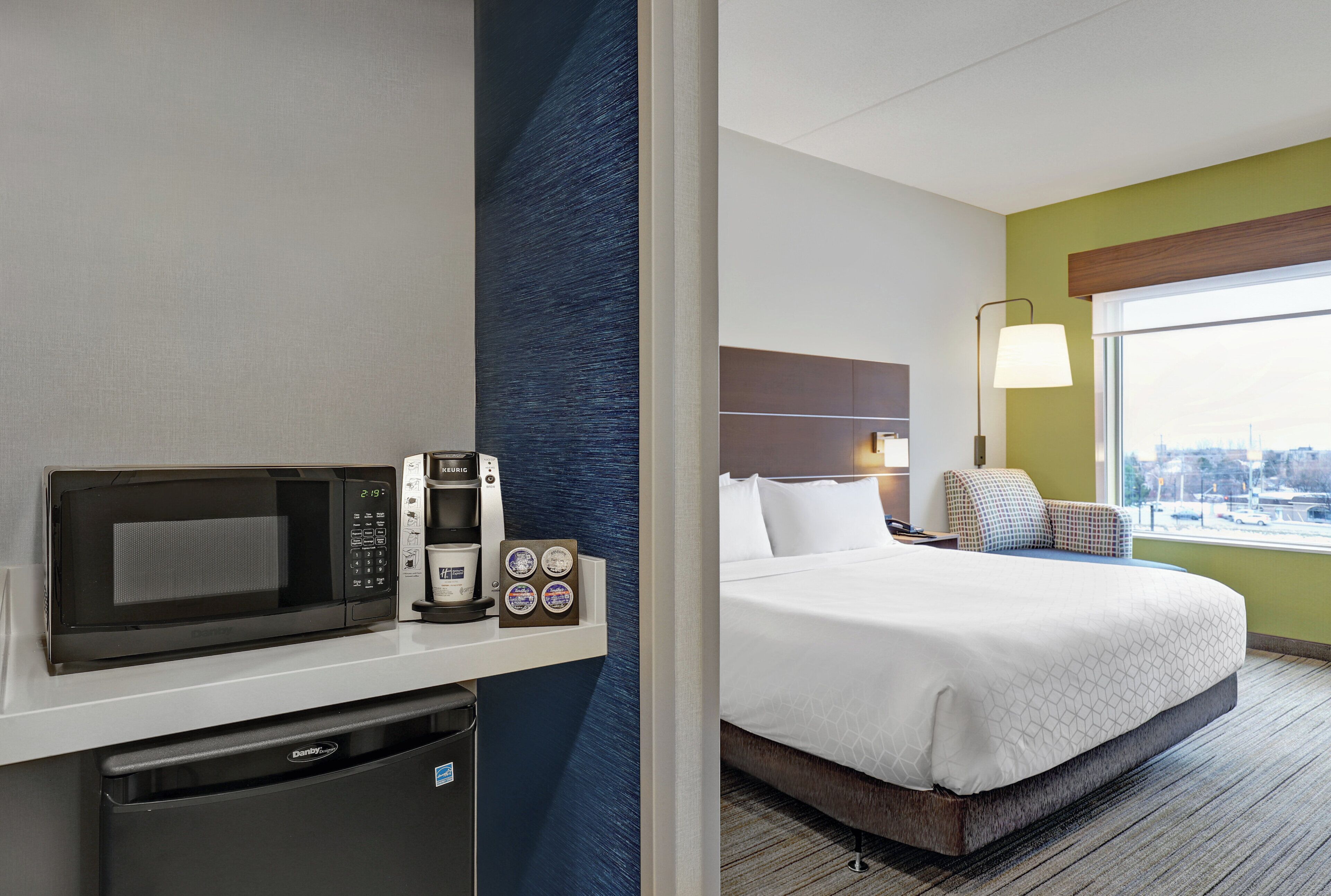 Habitación de hotel con cama, minibar, cafetera y vista a la ciudad.