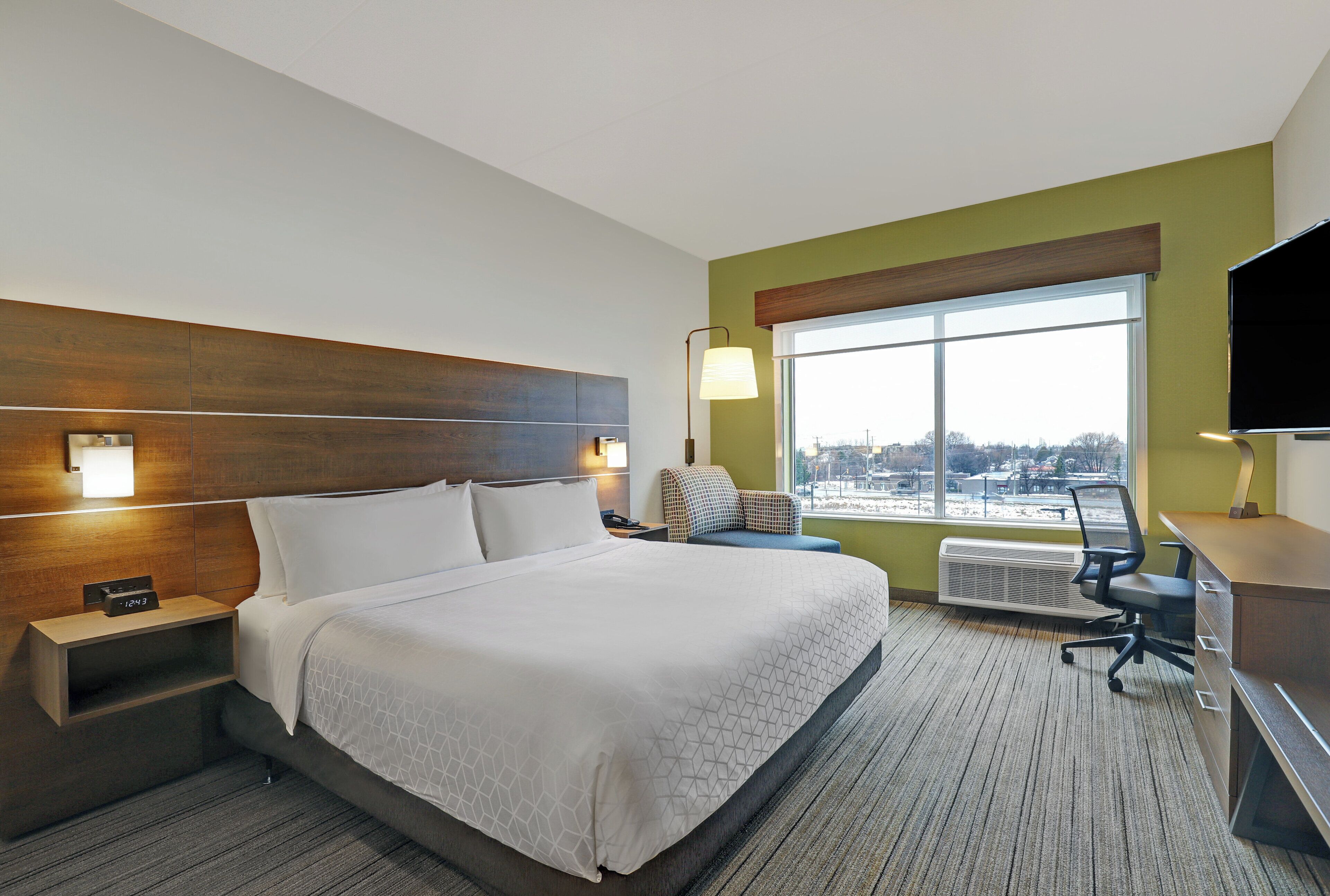 A hotel room with a large bed, a desk, a chair, a TV, and a window with a view of the city.
