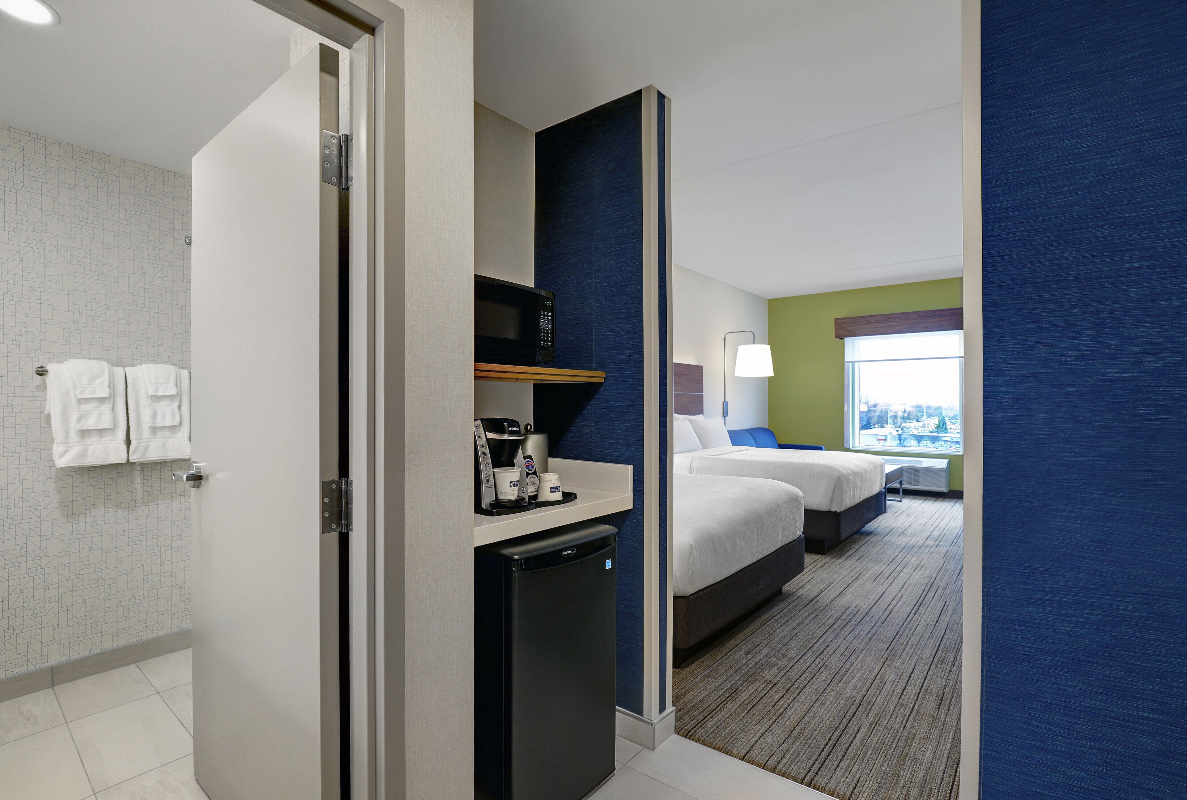 A hotel room with a bed, a desk with a coffee maker, a mini fridge, and a view of a cityscape through a window.