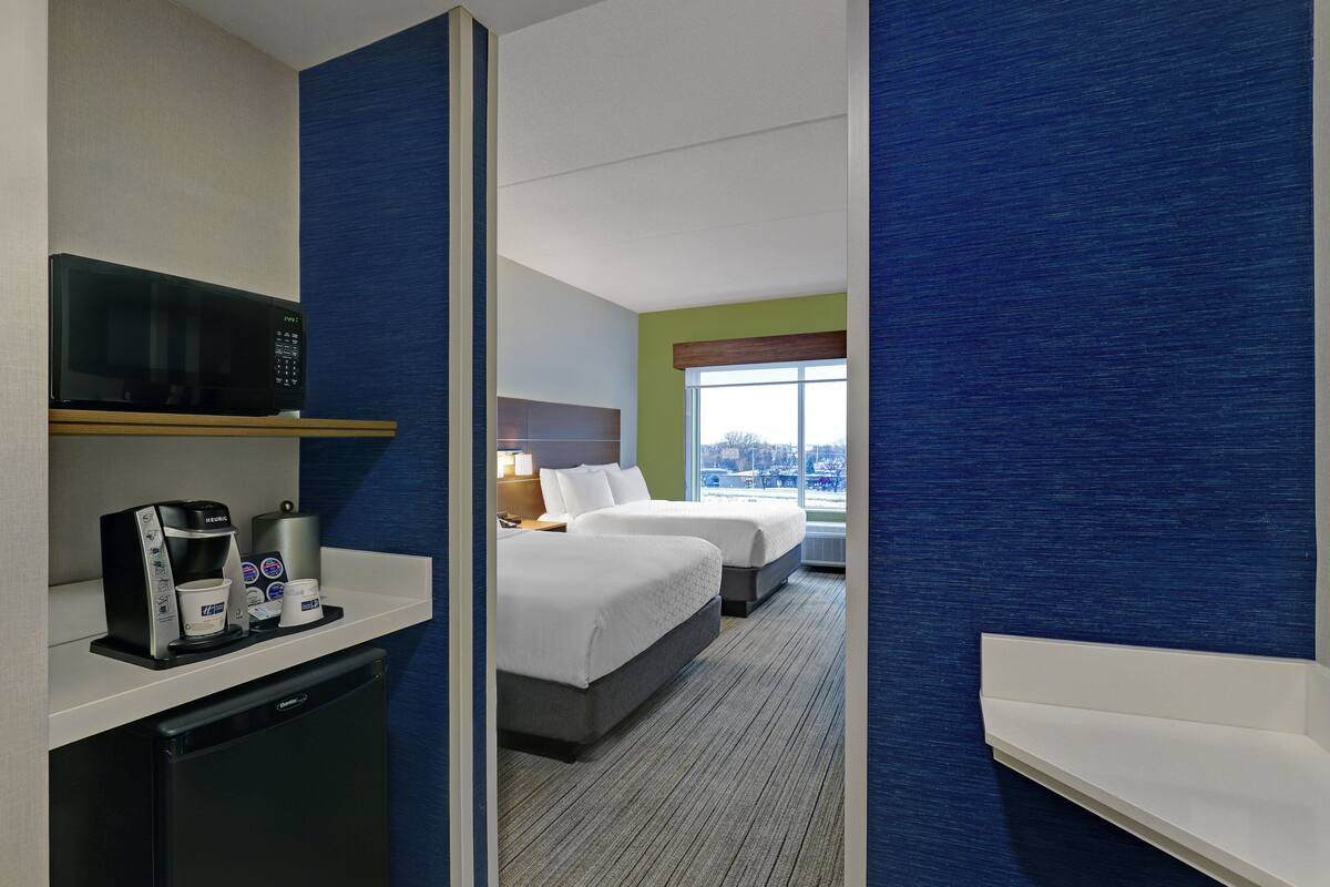 A hotel room with a kitchenette, two beds, a window with a view, and a blue accent wall.
