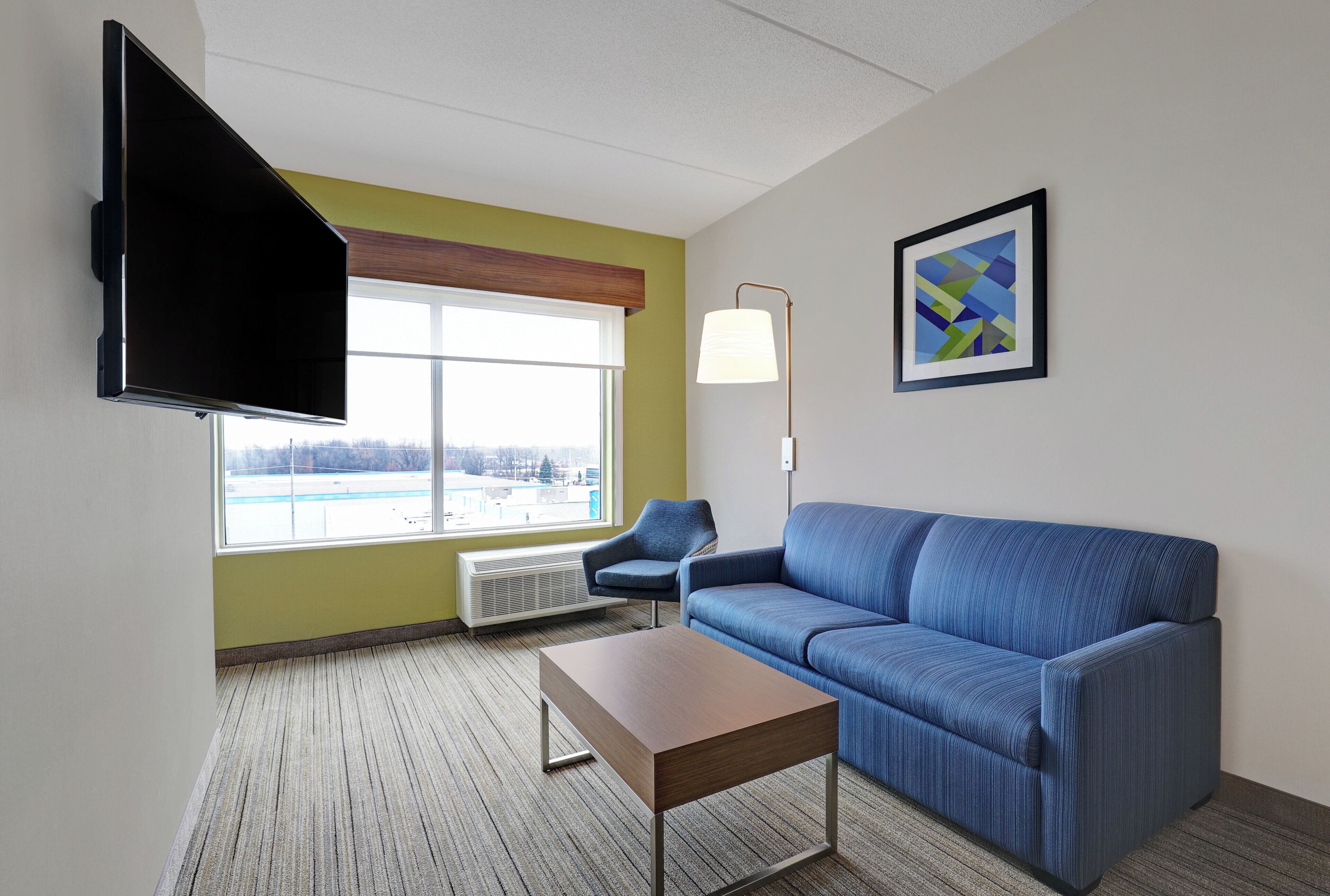 Une chambre d’hôtel moderne avec un canapé bleu, une table basse en bois, un téléviseur à écran plat fixé au mur, une lampe sur pied et une grande fenêtre donnant sur un immeuble.