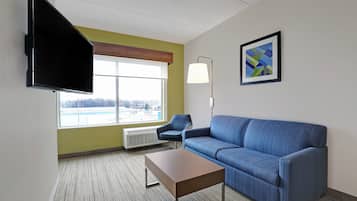 Une chambre d’hôtel moderne avec un canapé bleu, une table basse en bois, un téléviseur à écran plat fixé au mur, une lampe sur pied et une grande fenêtre donnant sur un immeuble.