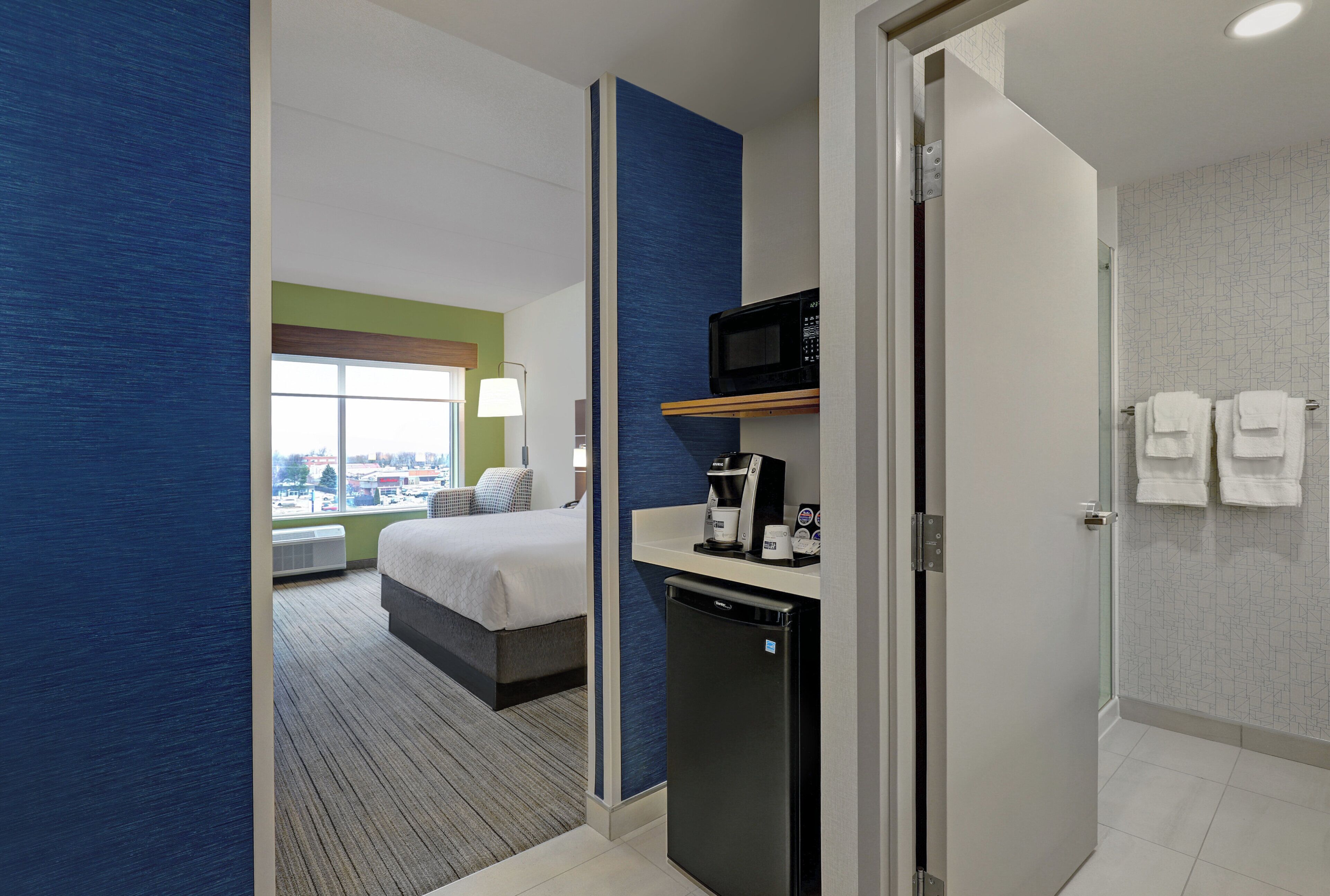 Habitación de hotel con cama, cafetera, microondas y baño con toallas.
