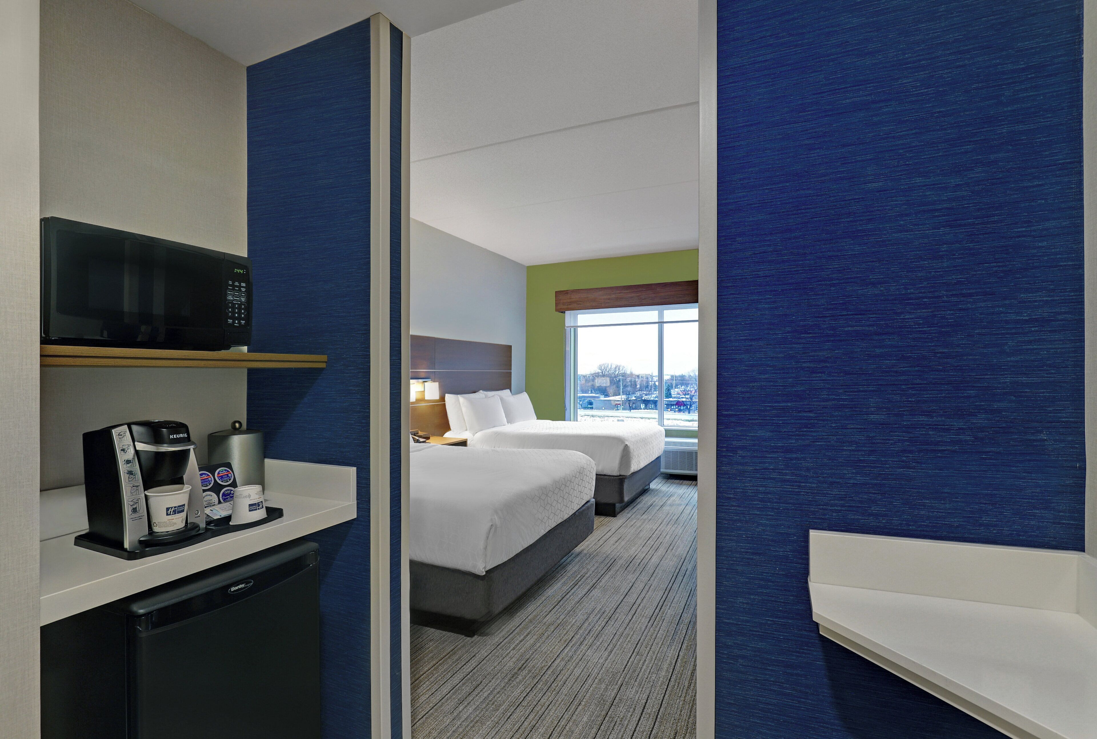 Habitación de hotel con kitchenette, una cama, ventanal con vistas y pared decorada en azul.