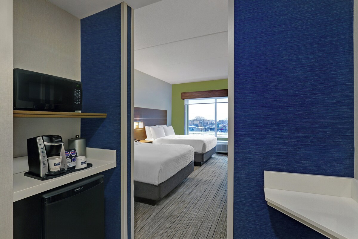 Une chambre d’hôtel avec une kitchenette, deux lits, une fenêtre donnant sur un paysage agréable et un mur d’accent bleu.