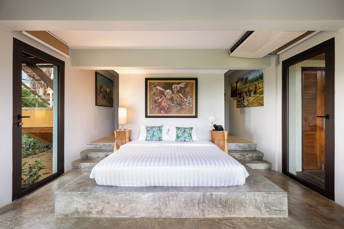 A bedroom with a large bed, a painting above the bed, and a view of a garden through a glass door.
