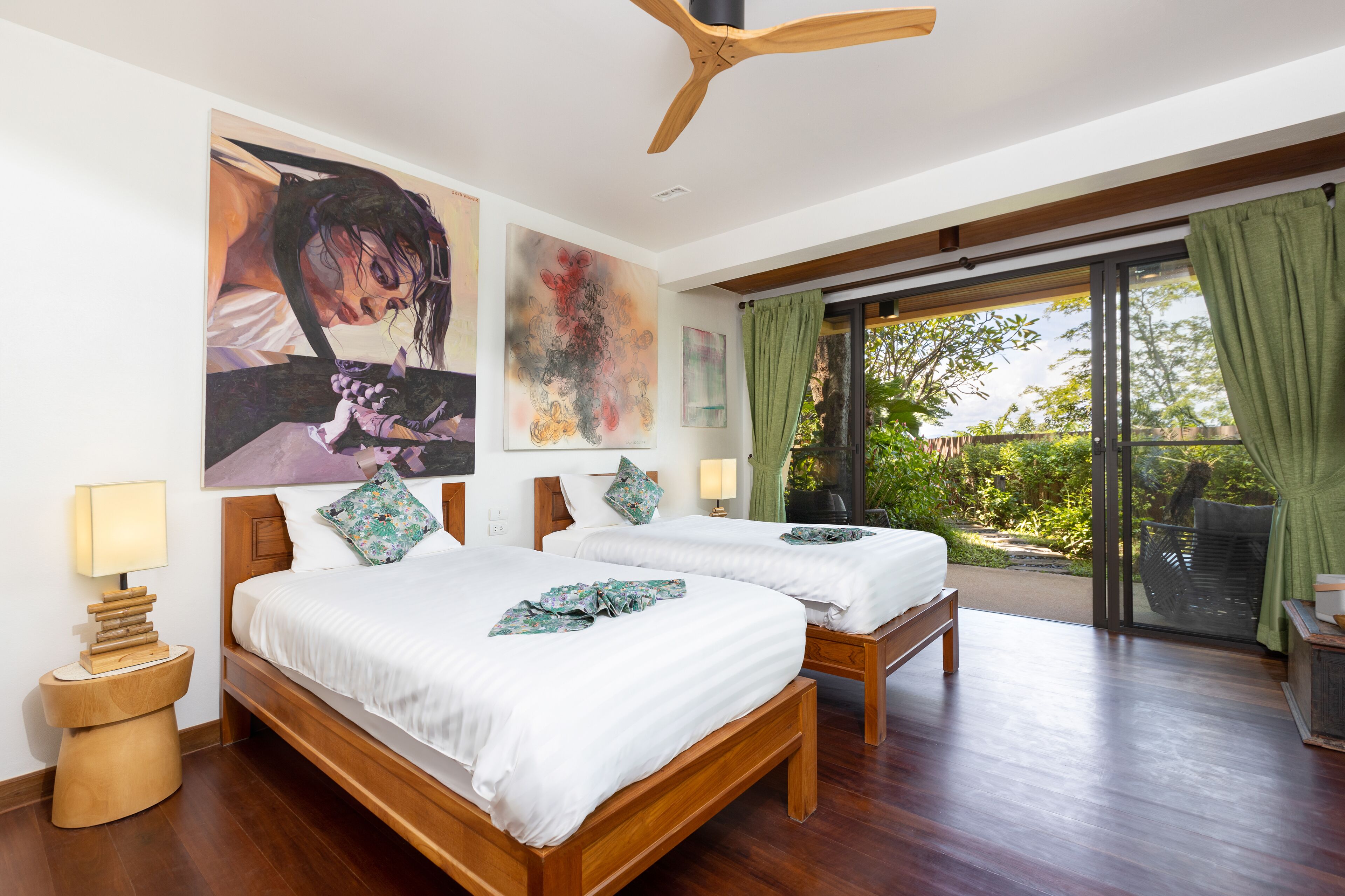 Quarto de hotel com duas camas, ventoinha de teto em madeira, janela ampla com vista para o verde e obras de arte nas paredes.