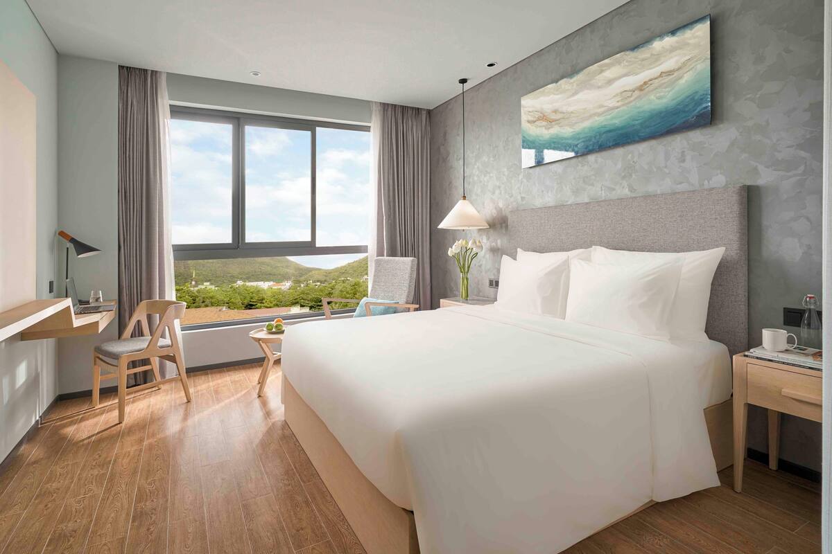 A hotel room with a large bed, a desk, a chair, a lamp, and a view of the outdoors.
