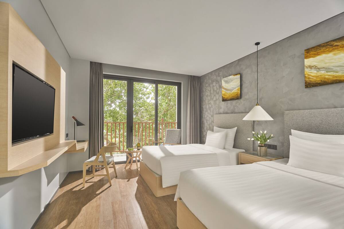 A modern hotel room with two beds, a flat-screen TV, a balcony with a table and chairs, and artwork on the walls.