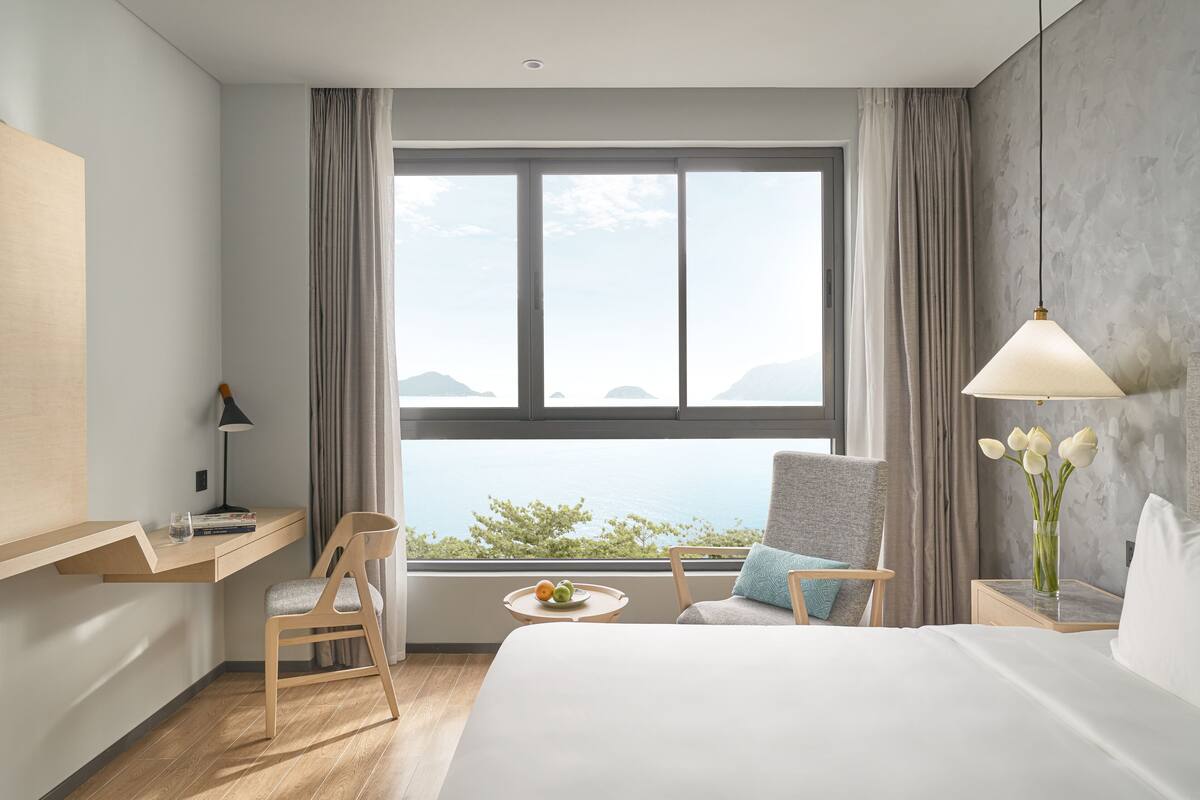 A hotel room with a large window, a bed, a desk with a lamp, a chair, a small table with fruit, and a vase with flowers.