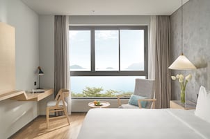 Ein Hotelzimmer mit großem Fenster, einem Bett, einem Schreibtisch mit Lampe, einem Sessel und einem kleinen Tisch mit Früchten sowie einer Vase mit Blumen.
