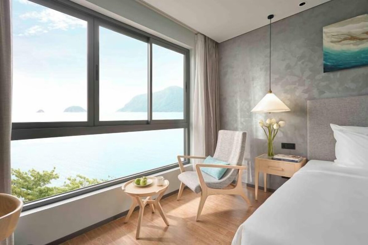 Habitación de hotel con un ventanal grande, una cama, una silla, una mesita y una mesita de noche.